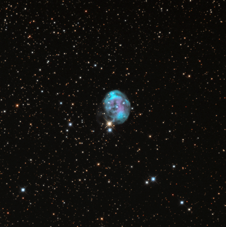 A colorful planetary nebula, NGC 7008 (also known as the Fetus Nebula), glows in shades of turquoise, pink, and purple against a dense star field. The nebula appears roughly spherical with wispy, complex internal structures. Bright foreground stars, including one with prominent diffraction spikes, add contrast to the dark background of deep space.